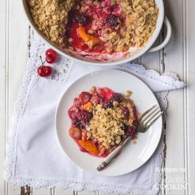 An overhead photo of a serving of summer fruit crisp on a plate with a fork and the entire pan behind it.